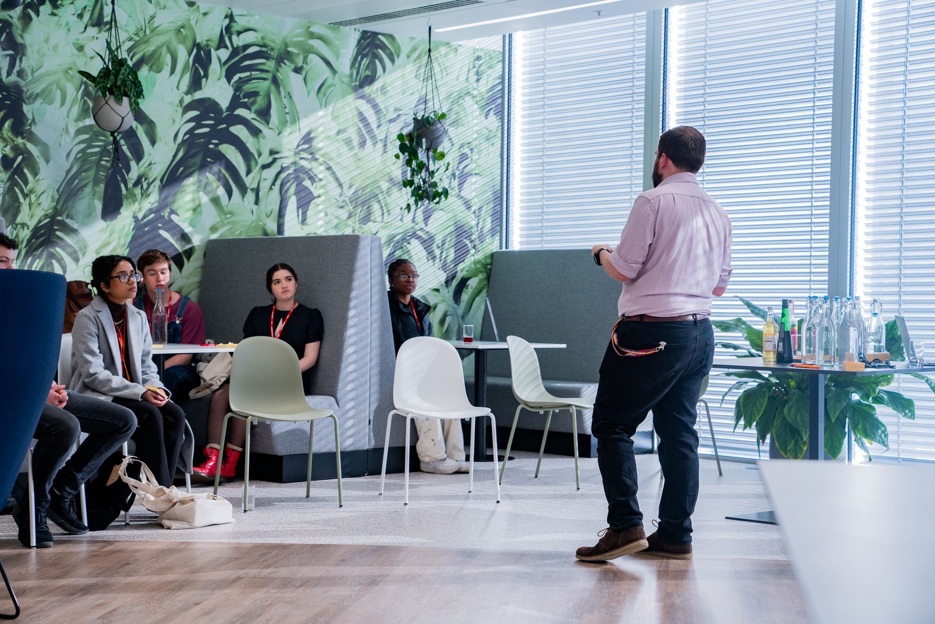 Presenter speaking to seated audience in a bright modern room with large windows and plants