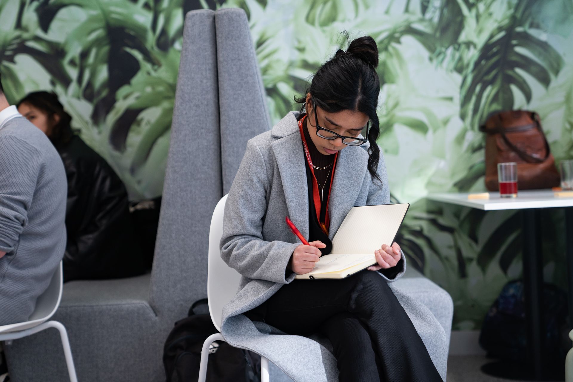 Woman in a gray coat reading a document while seated in a lounge with green wallpaper.