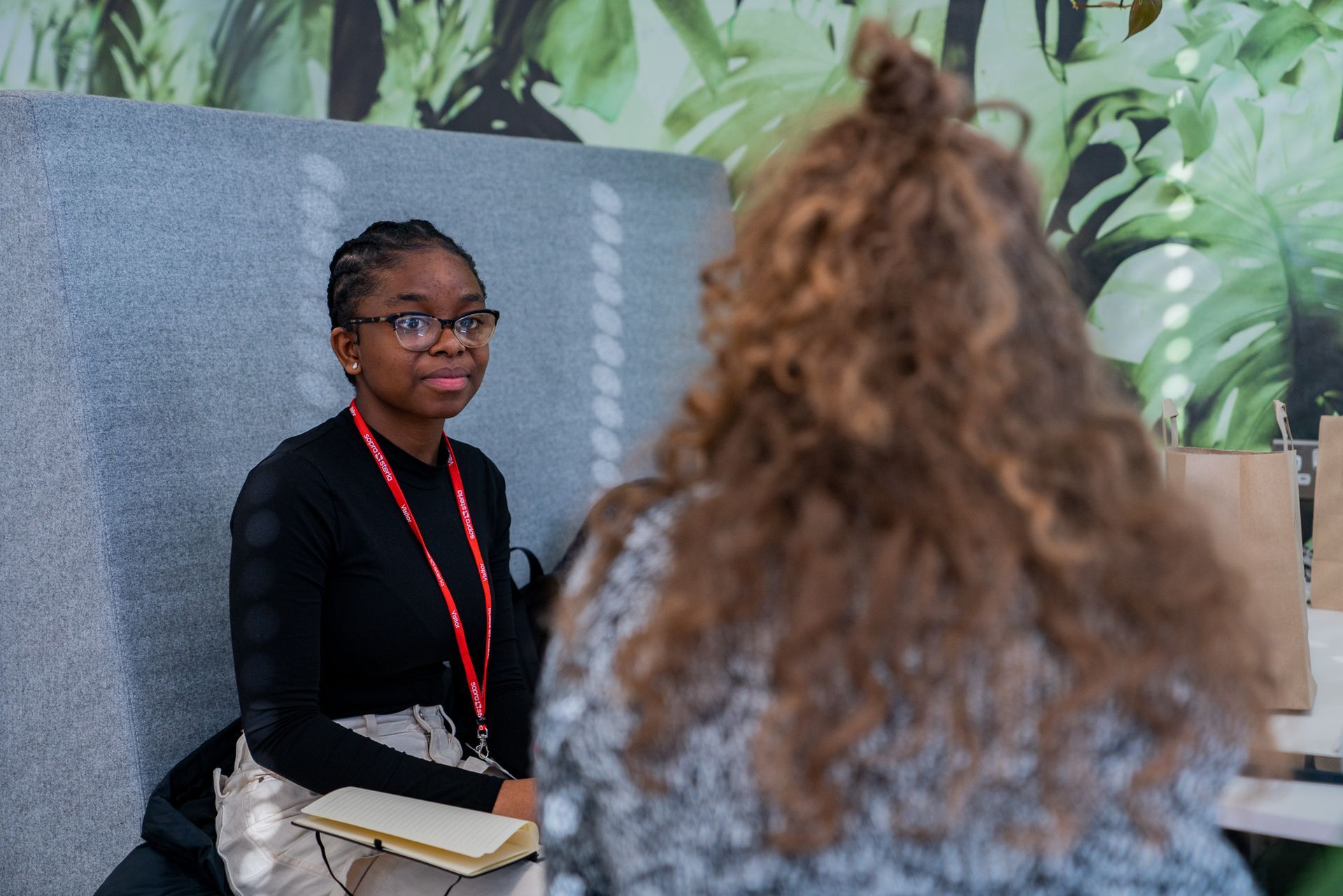 Two people seated in conversation at a table, with one facing the camera and a leafy mural behind them.