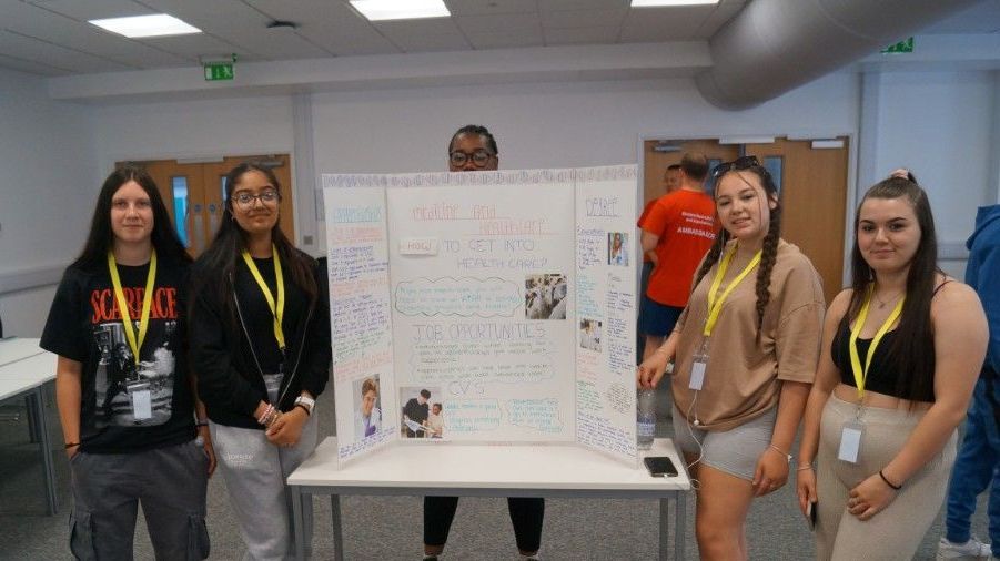 Five people stand around a poster board presentation in a classroom or conference room.