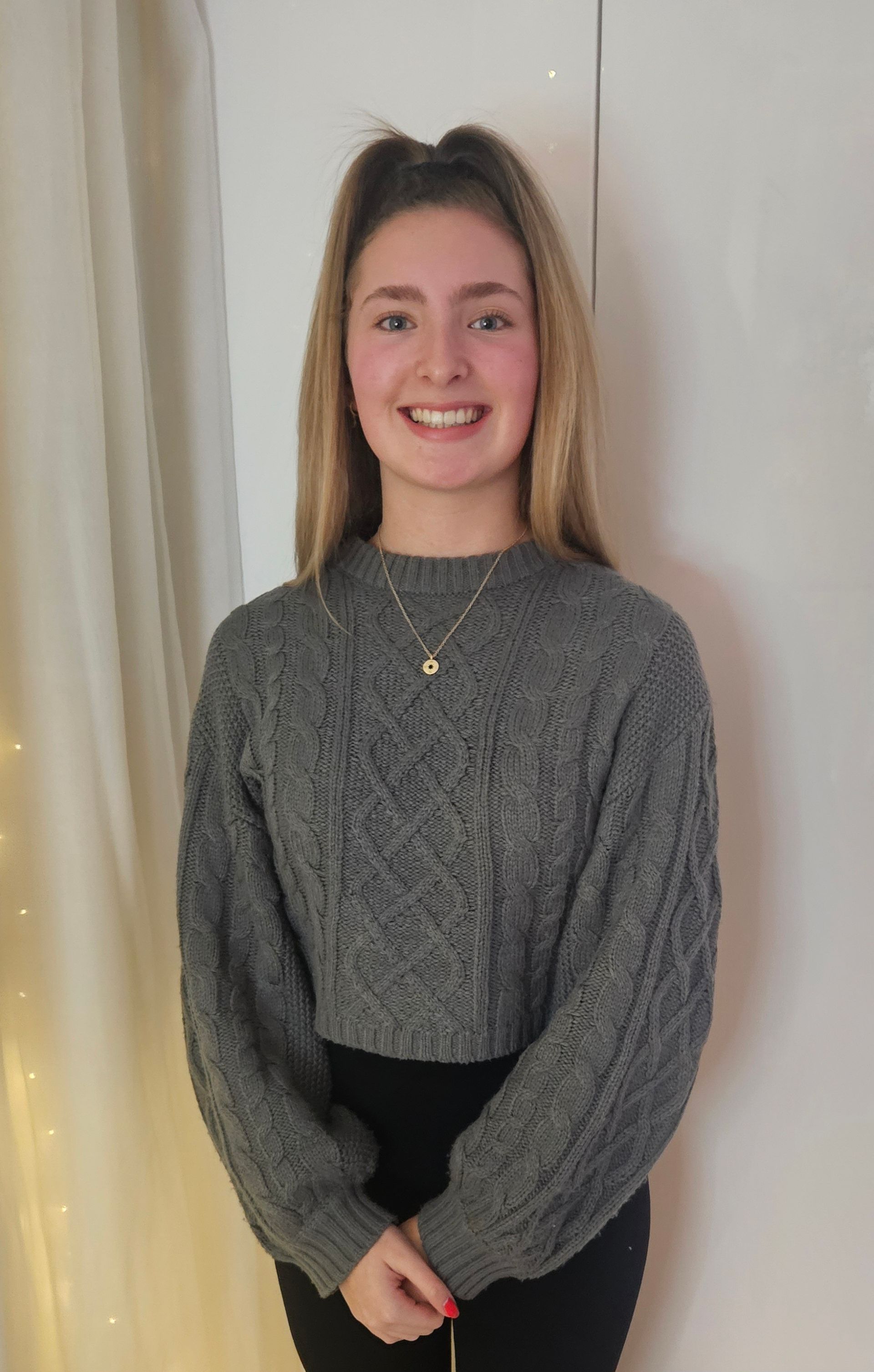 Smiling person in a gray sweater standing indoors against a light curtain backdrop