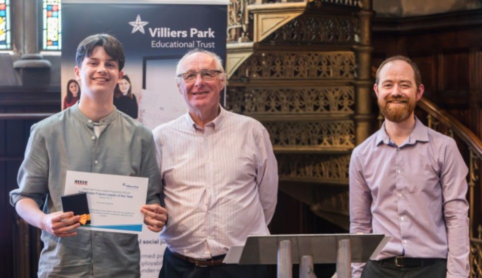 Three men stand on stage, one holding a certificate, with a Villiers Park banner behind them.