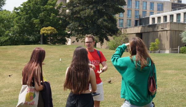 Three people standing on a grassy lawn talking near a building, one holding a phone up.