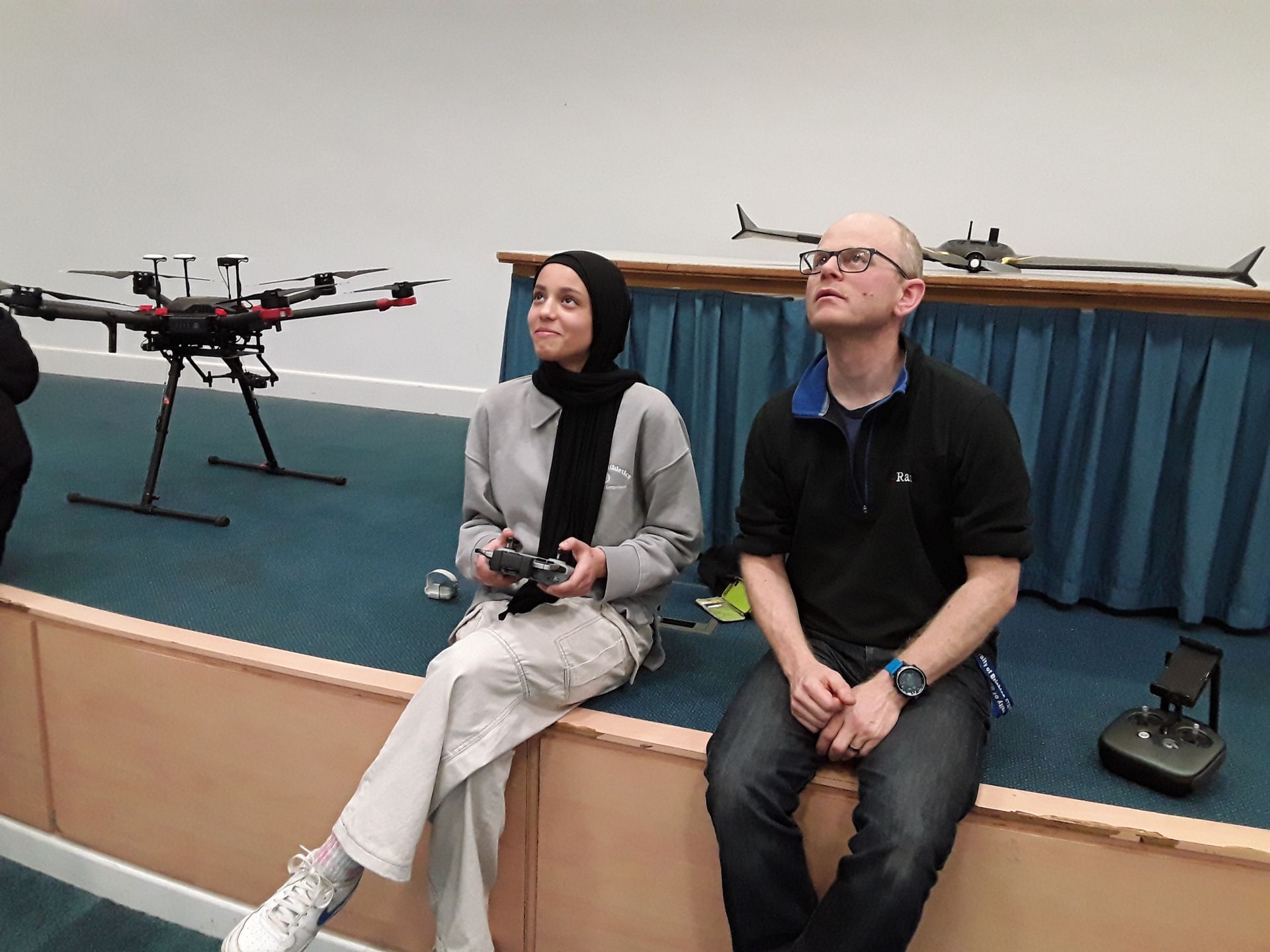 Two people sitting on a stage, one holding a remote controller, with drones displayed behind them.