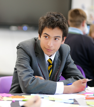 Student in a gray blazer and striped tie writing at a table in a classroom.