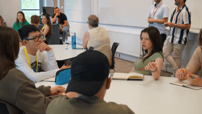 People in a bright meeting room gathered around a table in discussion, with notebooks and laptops.