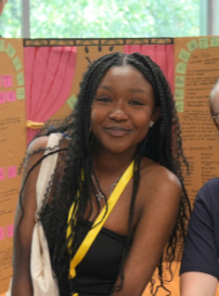 Smiling person with long braids and yellow lanyard at an indoor event booth