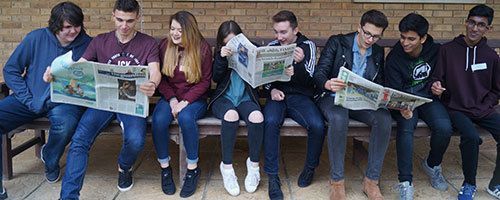 Group of students sitting on a bench, reading newspapers and smiling outdoors