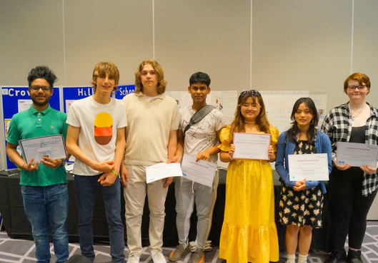 Seven students standing indoors, several holding certificates in front of a banner.