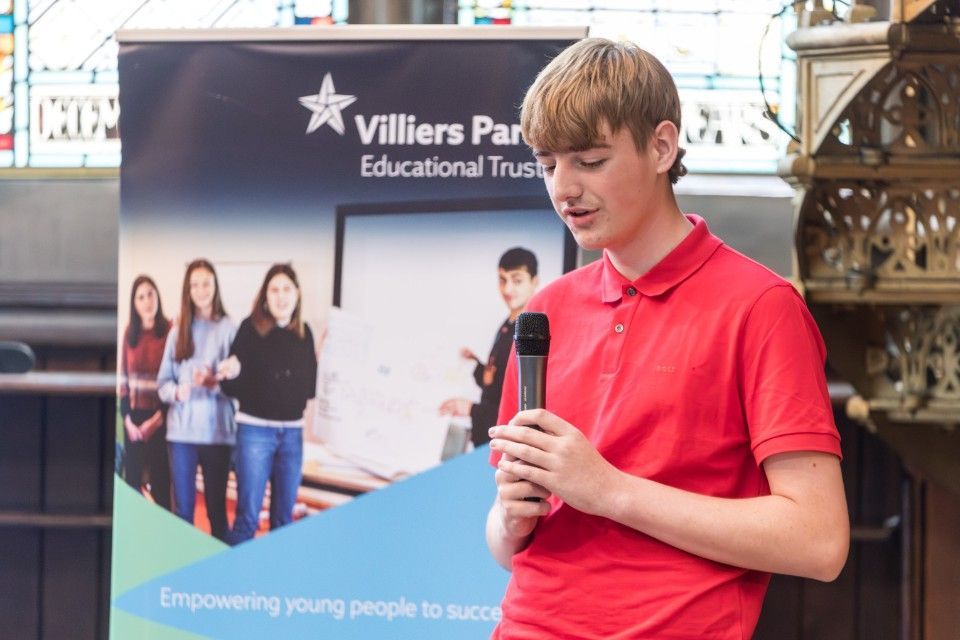 Young man in a red shirt speaking into a microphone at a Villiers Park Educational Trust event.