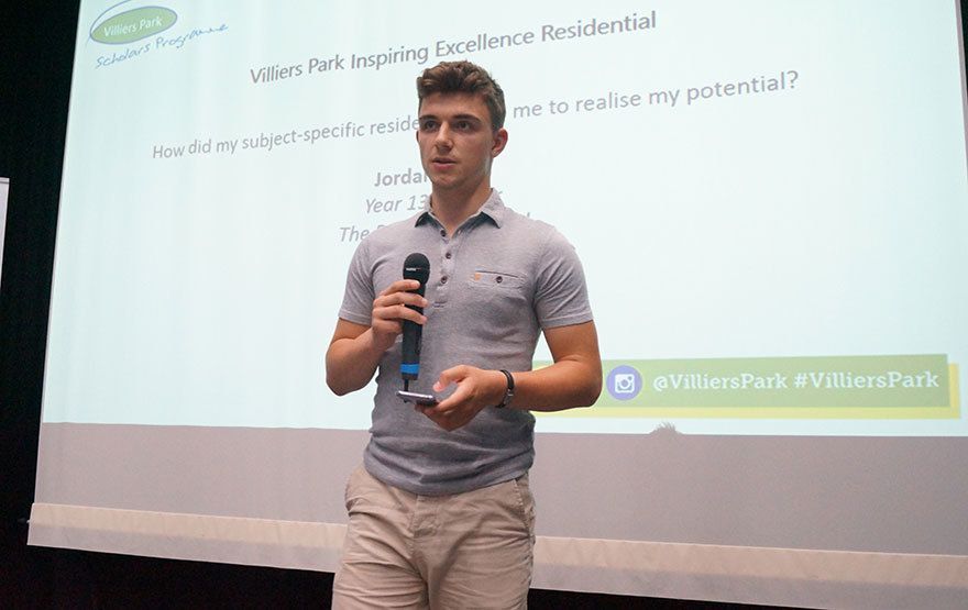 Man speaking into a microphone in front of a projected presentation slide at an indoor event