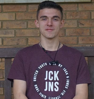 Young man in a maroon JCK JNS T-shirt standing against a brick wall, smiling softly.