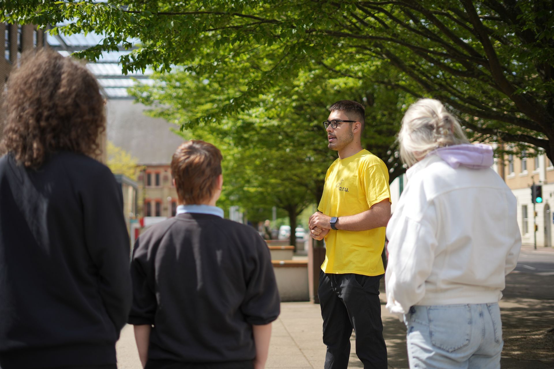 Small group listening to a man in a yellow shirt speaking outdoors under leafy trees.