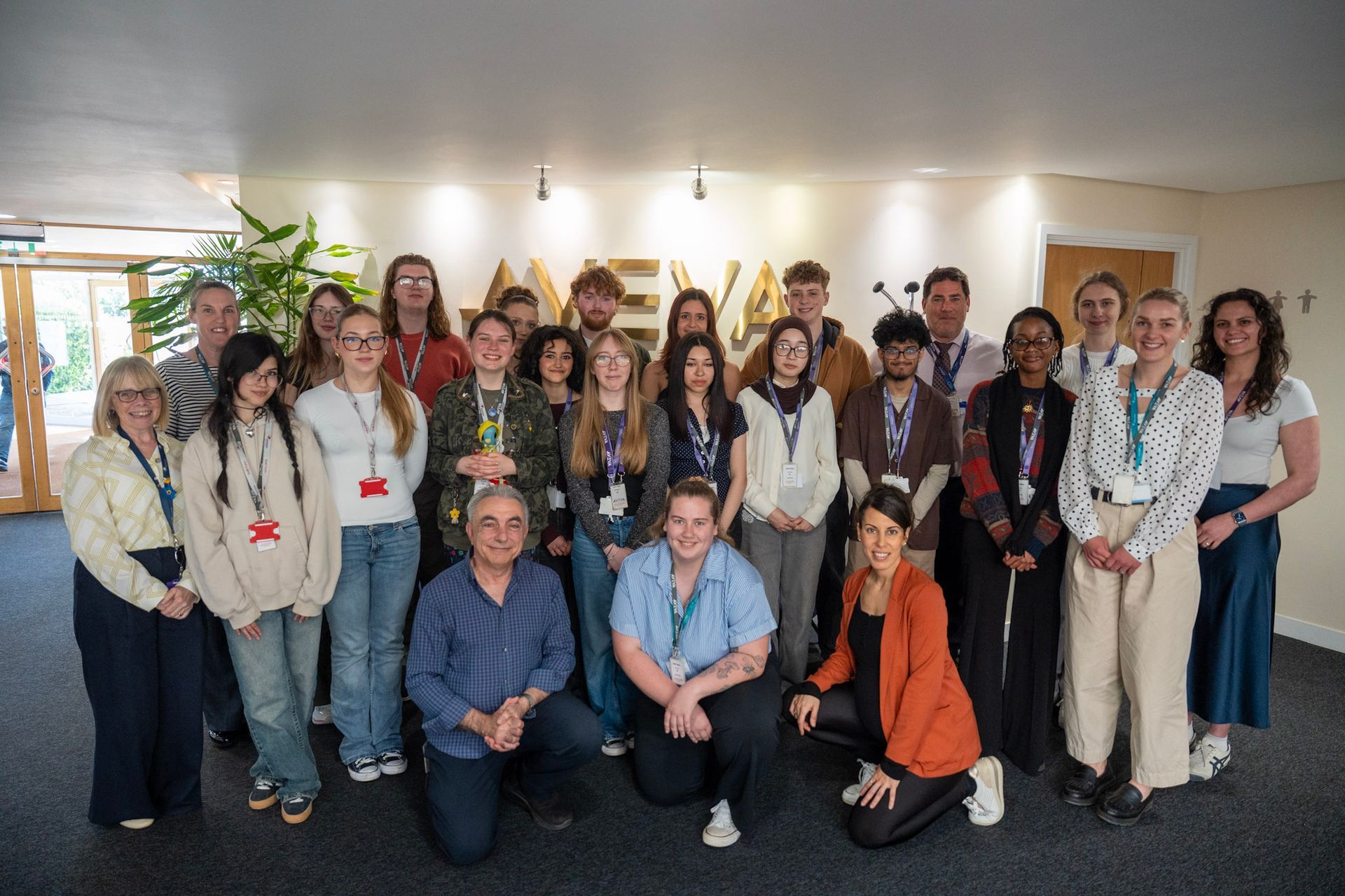 Large group of people posing indoors in front of a wall with plants, many wearing blue lanyards