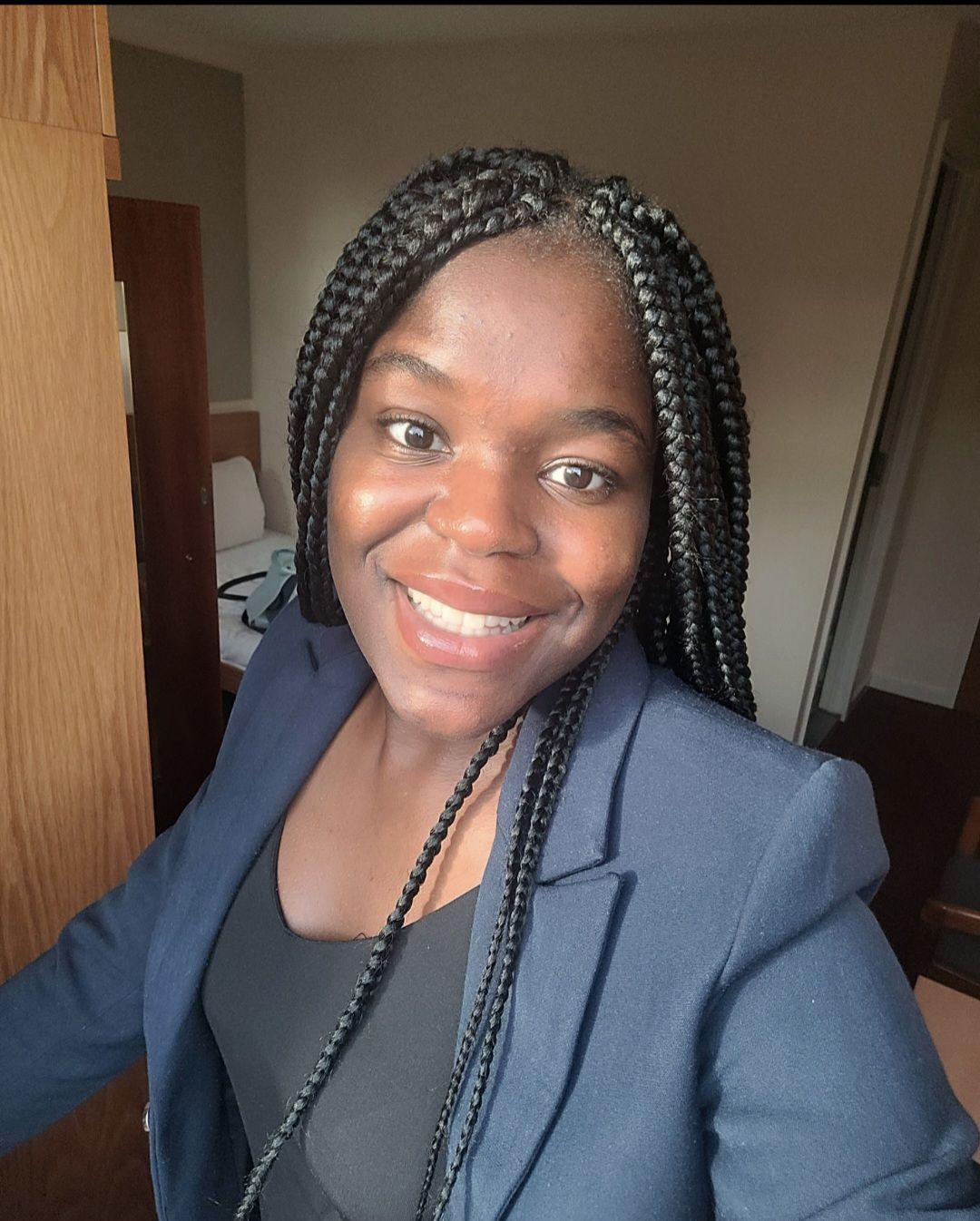 Smiling person with braided hair wearing a navy blazer indoors near a doorway.