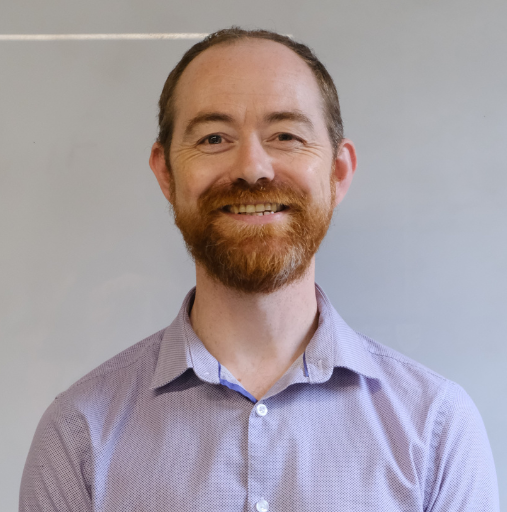 Smiling man in a light purple button-up shirt against a plain gray background