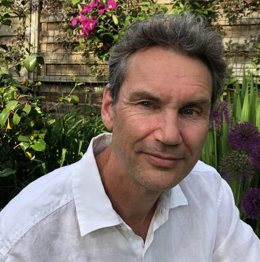 Man in a white shirt sitting in a garden with flowering plants and stone wall background