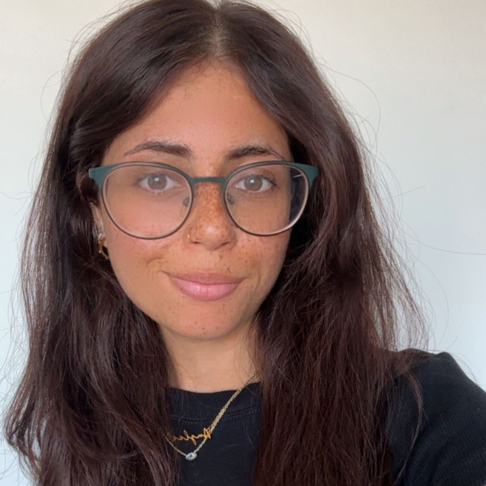 Woman with glasses and long dark hair, wearing a black top and necklace against a plain light background.