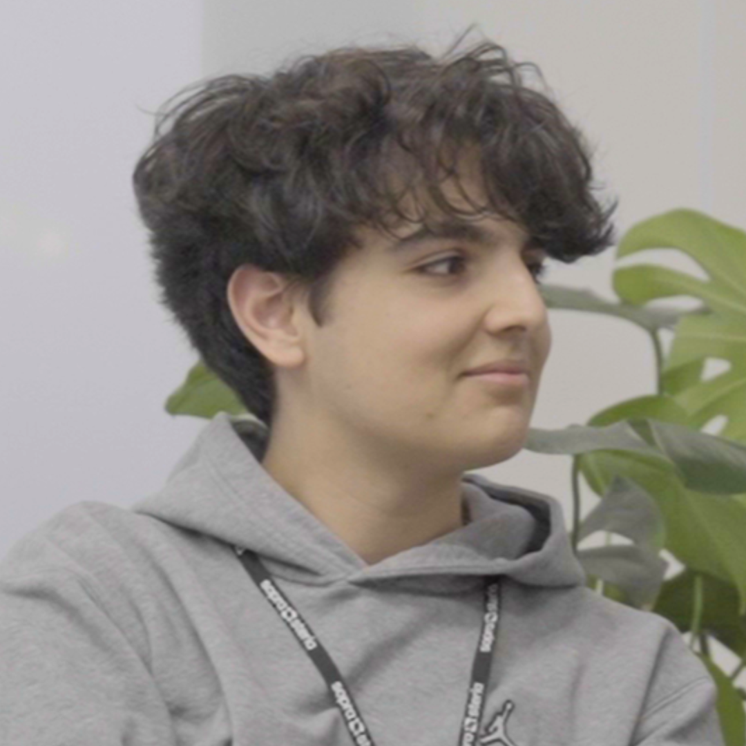 Person in a gray hoodie with curly dark hair, seated indoors beside a green plant, looking to the side.