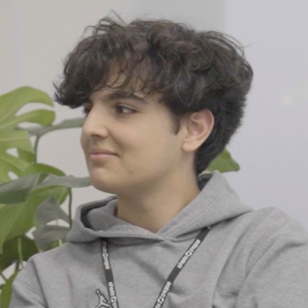 Person in a gray hoodie looking left, seated indoors beside a green plant.