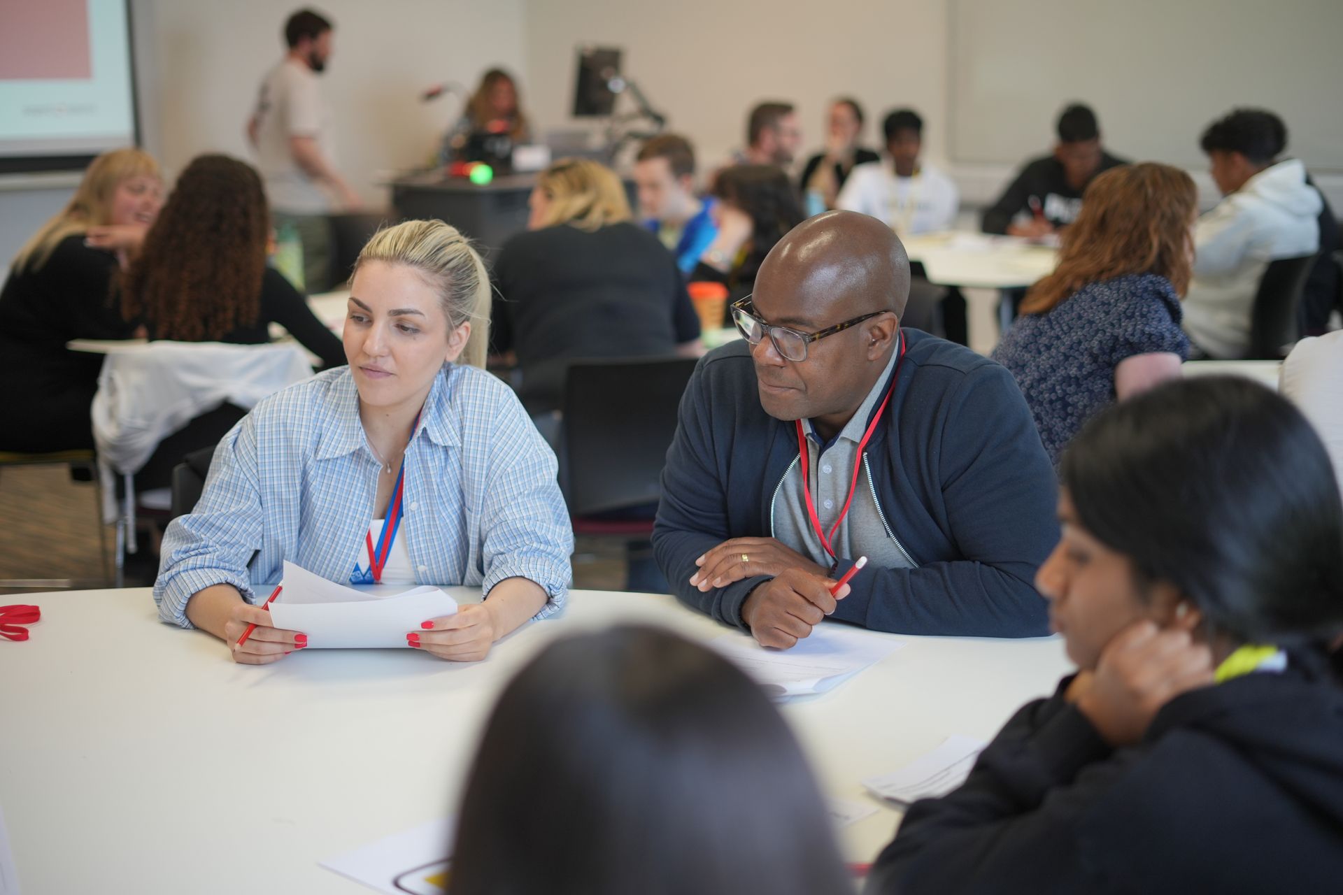 People seated at tables in a workshop, discussing papers in a bright classroom.
