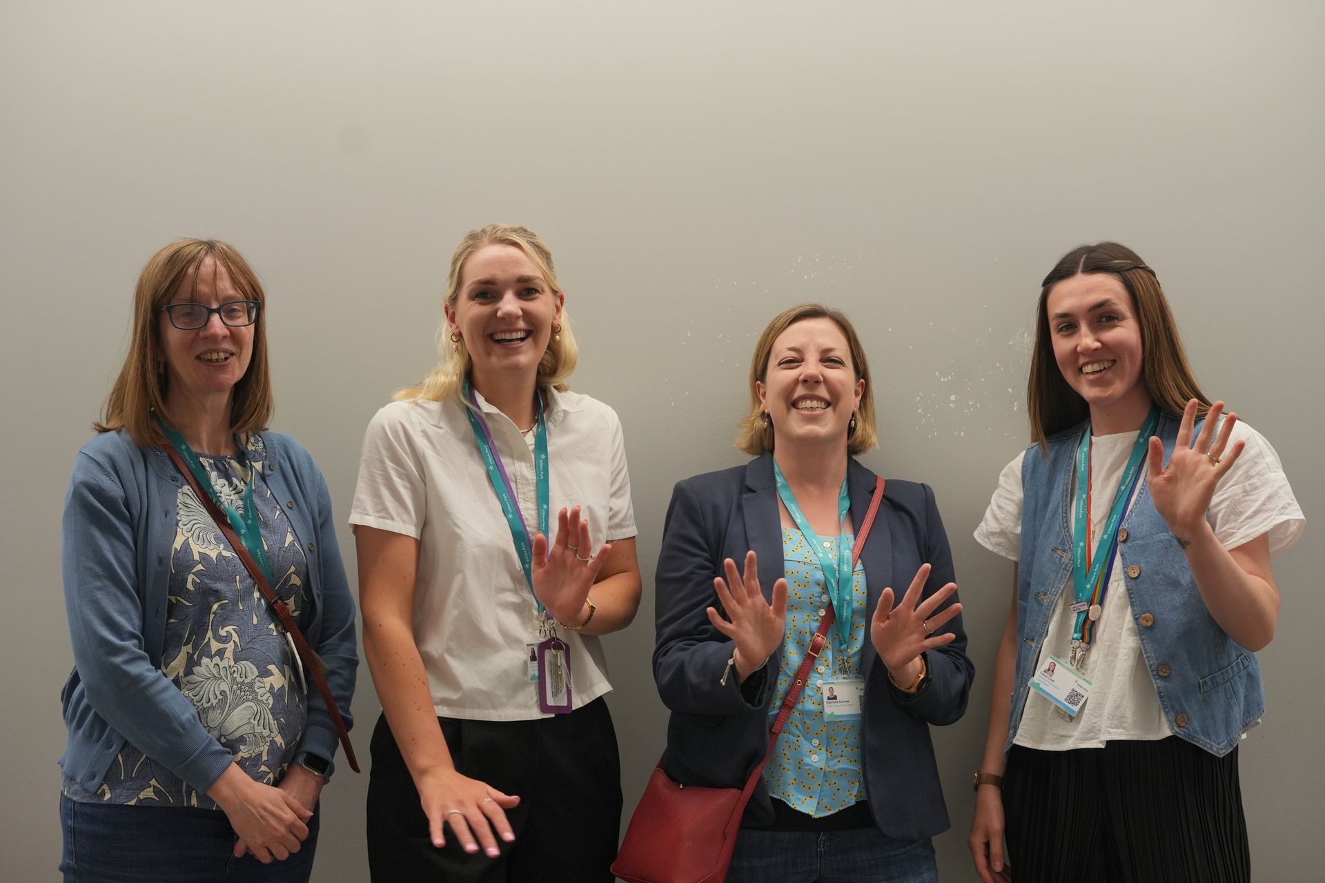 Four smiling people stand indoors, wearing lanyards and waving at the camera.