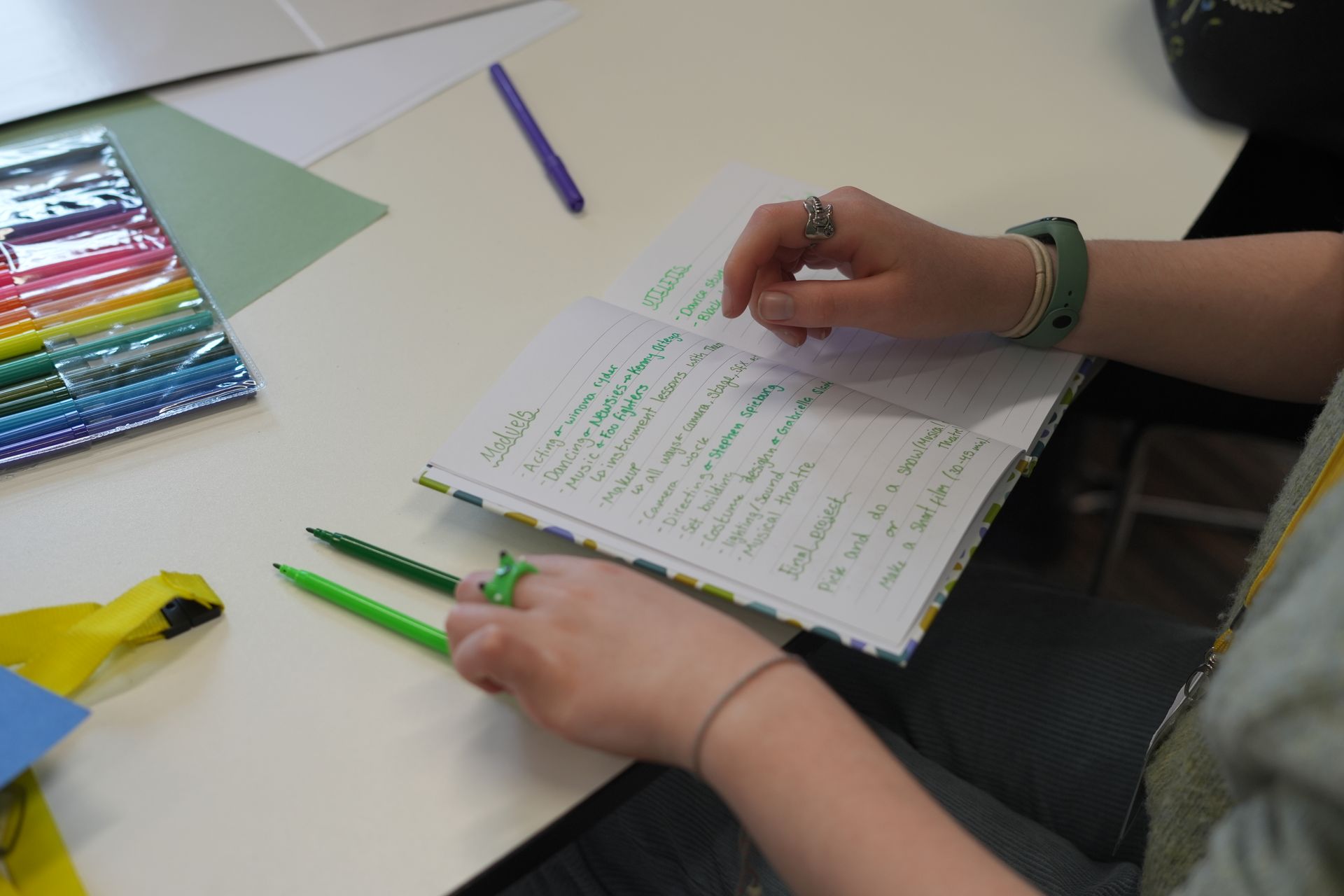 Hands writing notes at a desk with green pens and colored pencils nearby