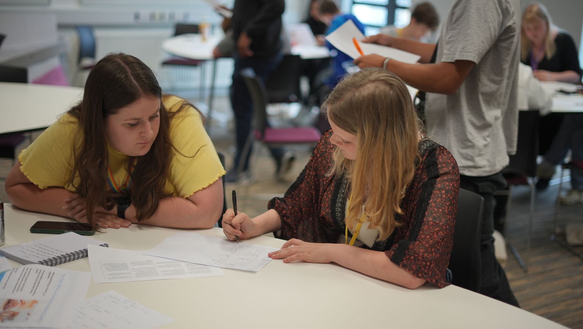 Two students collaborating at a table in a classroom, writing on paper with other students in the background