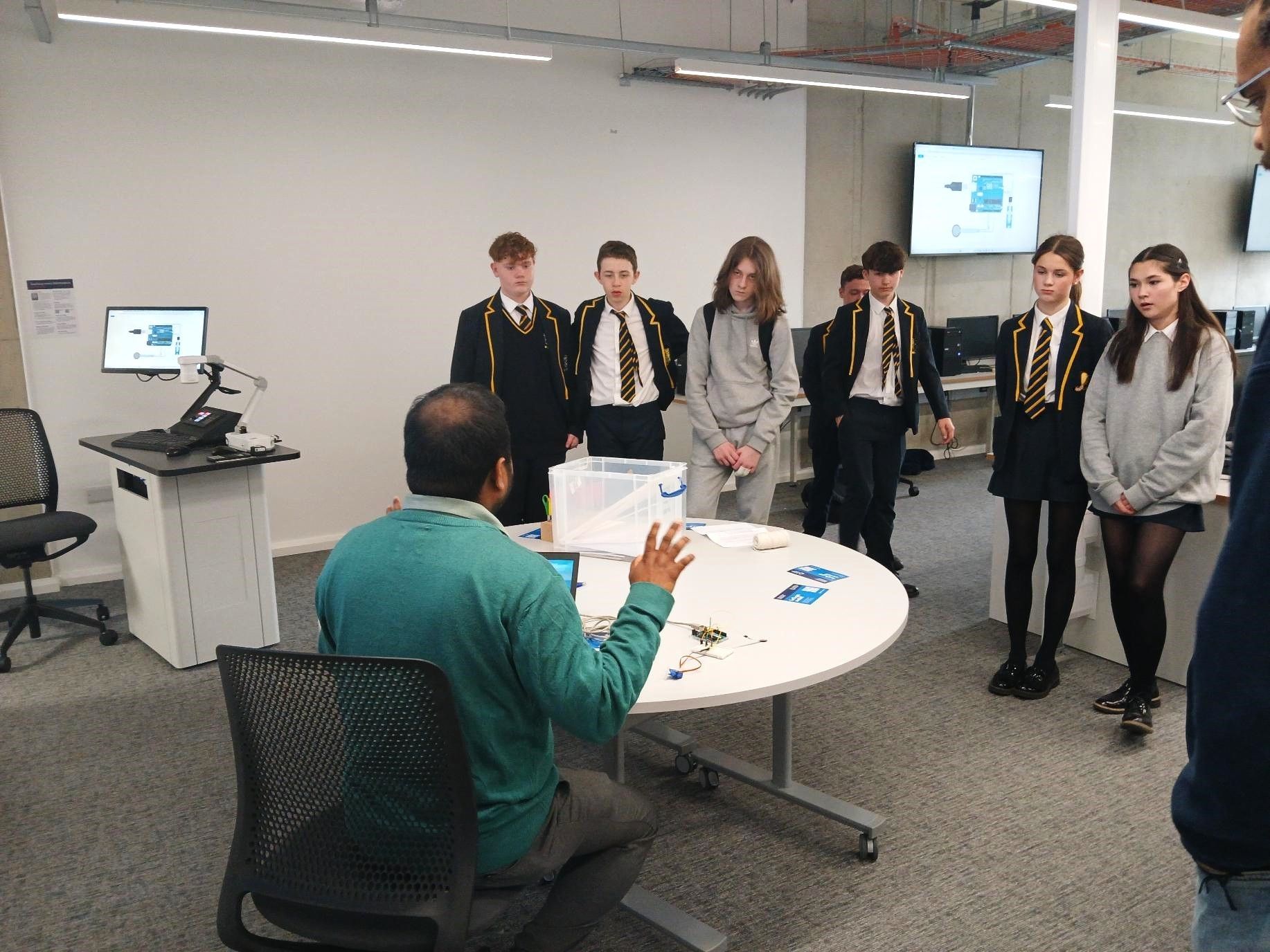Teacher presenting to a small group of students in a classroom with a projector and round table.