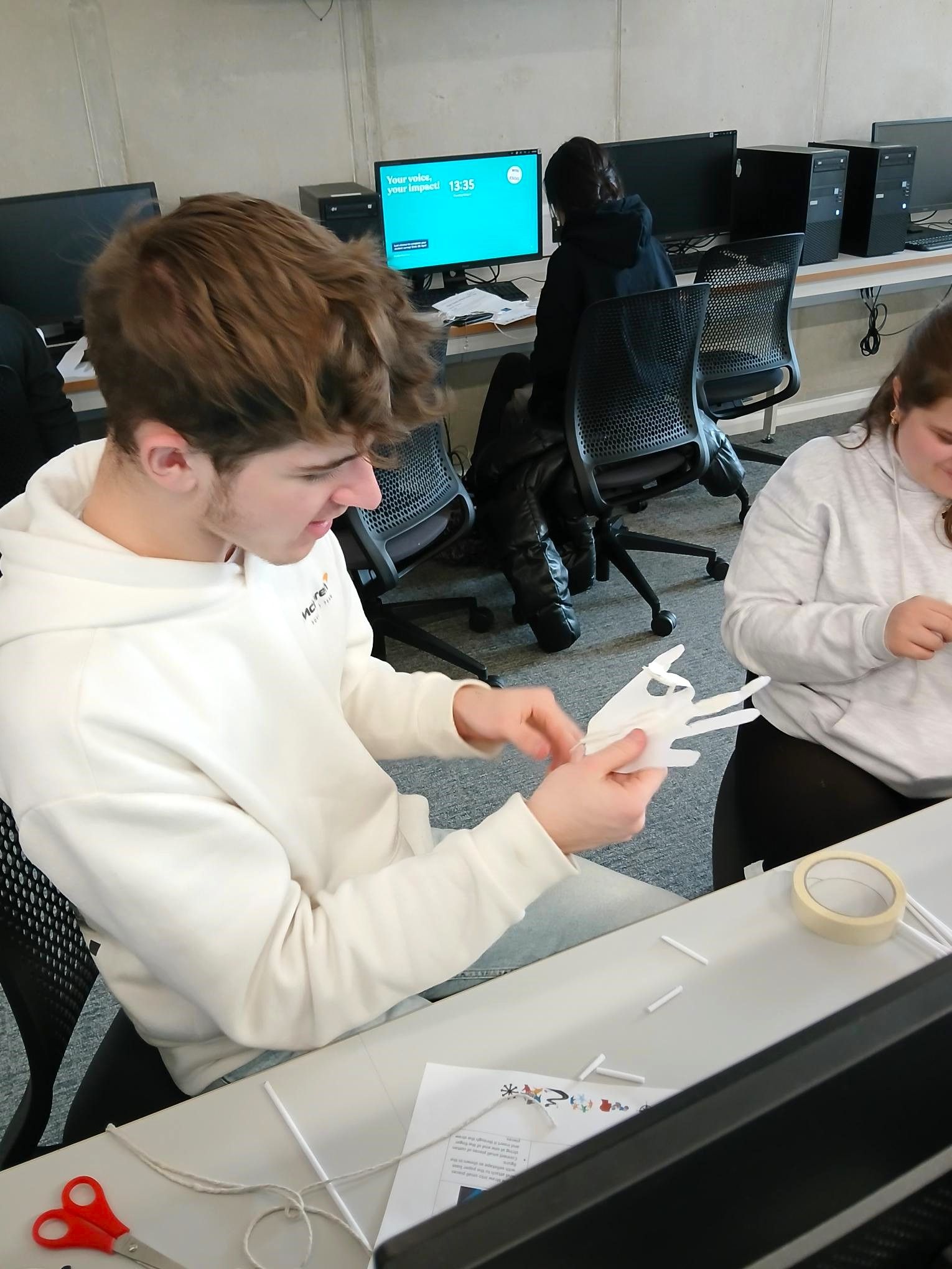 Person in a white hoodie assembling a white paper object at a desk in a computer lab