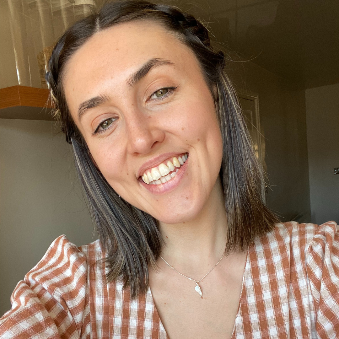Smiling person in a brown plaid shirt, selfie indoors near a sunlit window.