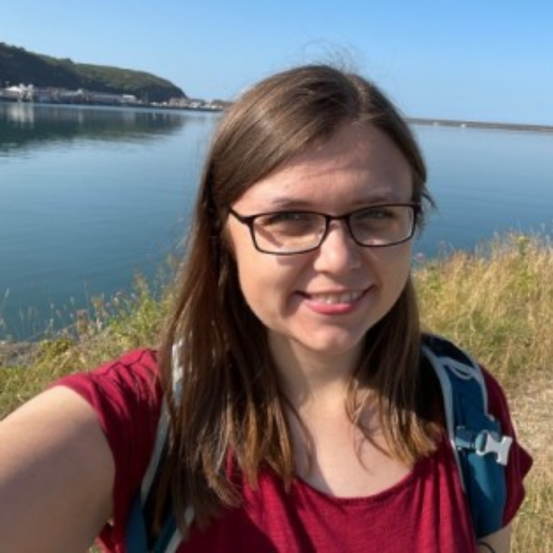 Woman smiling for a selfie by a calm lake on a sunny day, wearing glasses and a red shirt.