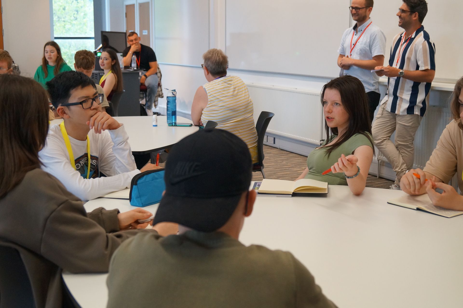 People seated at tables in a bright meeting room, talking and listening during a group discussion.