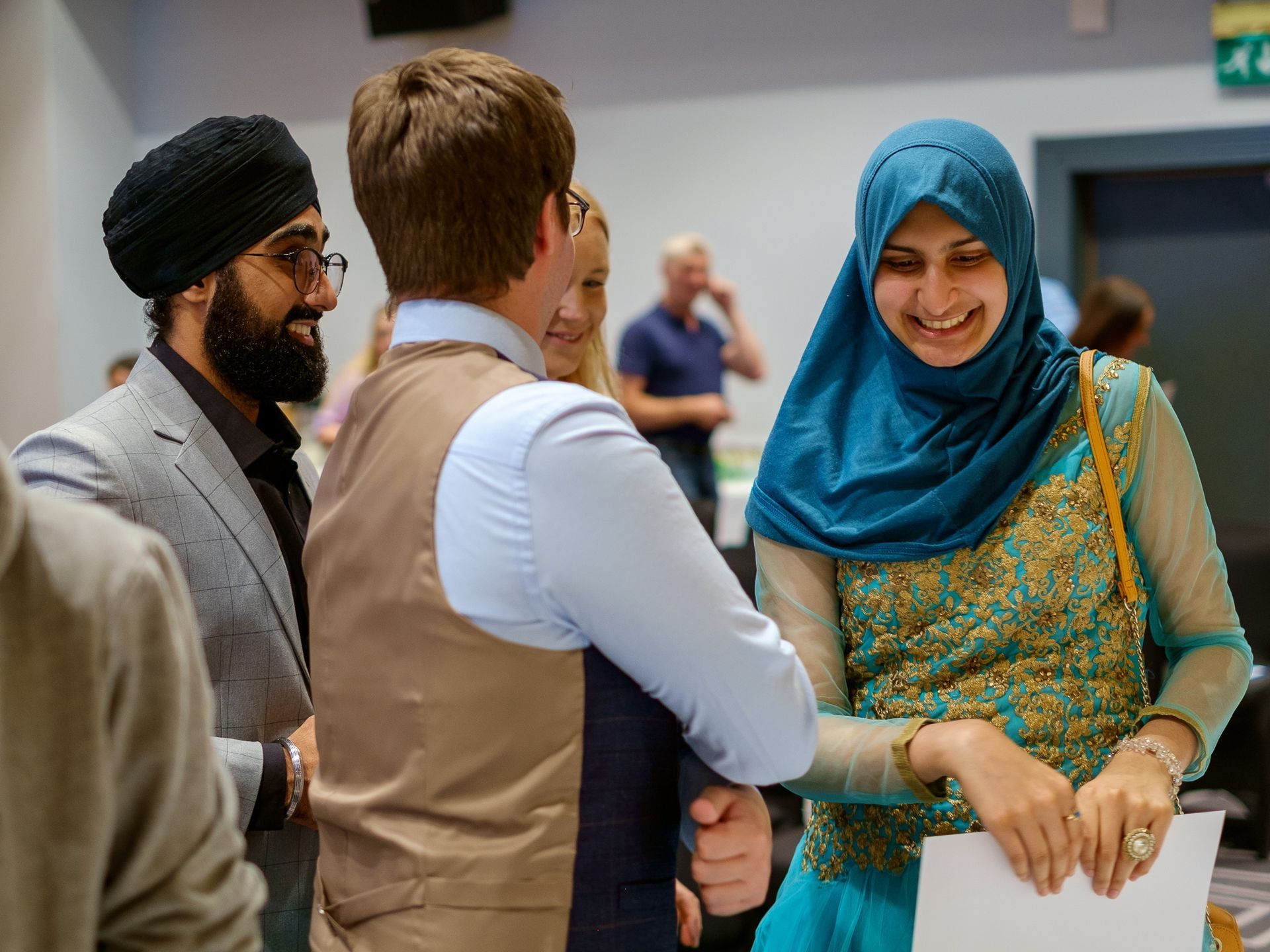 Three people talking at an indoor event, with one woman in a teal headscarf smiling.