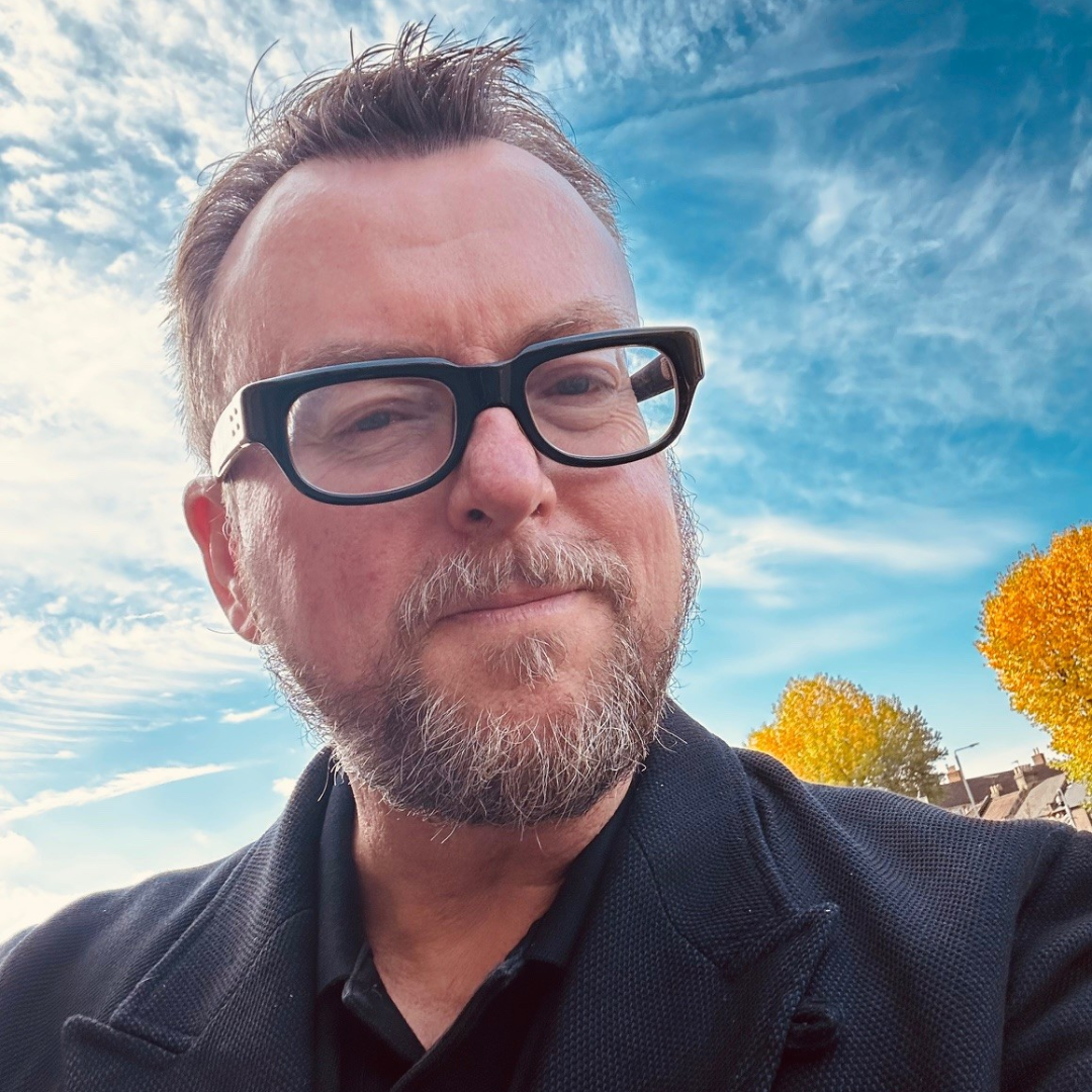 Man with glasses taking a selfie outdoors under a blue sky with autumn trees behind him