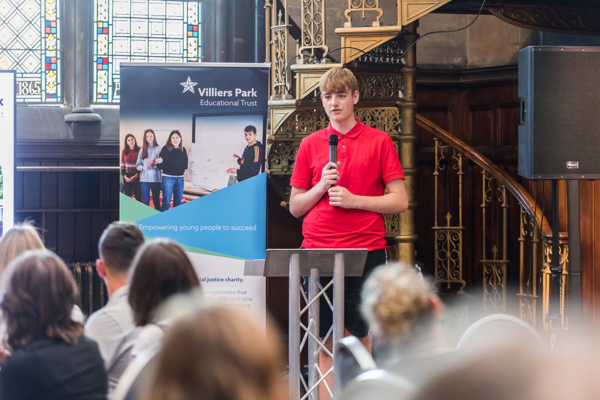 Speaker in red shirt addressing an audience at a podium in a church-like hall