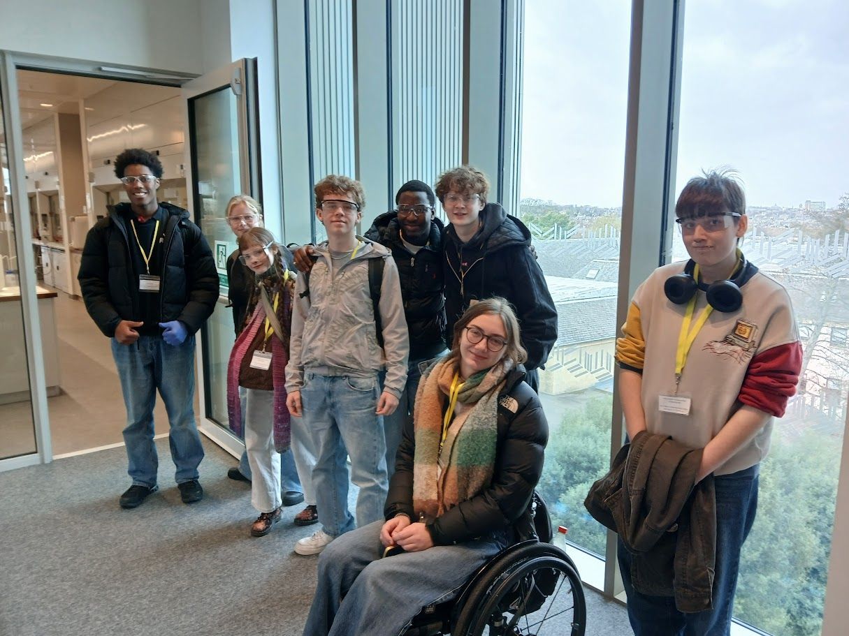 Group of six people standing indoors by large windows, one person seated in a wheelchair.