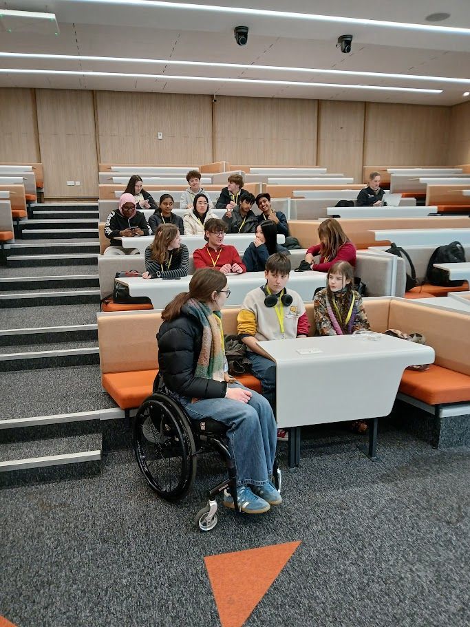 Students seated in a tiered classroom, discussing around tables; one person uses a wheelchair.