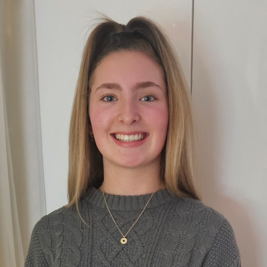 Portrait of a smiling person in a gray sweater and gold necklace, standing indoors against a light wall.
