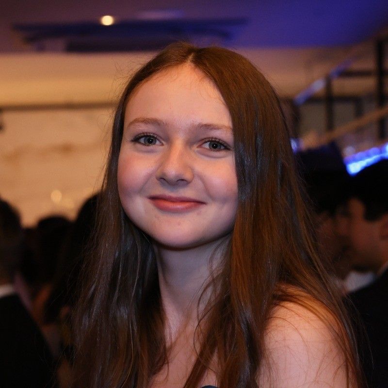 Smiling person with long brown hair at an indoor event with soft purple lighting
