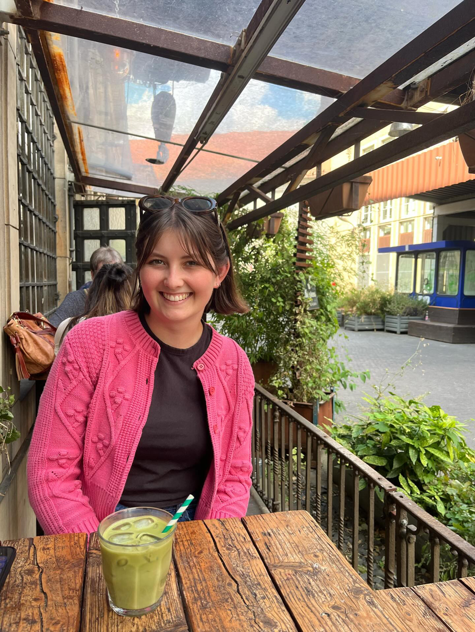 Smiling woman in a pink sweater sits at a wooden table in a covered outdoor café, with a green drink.