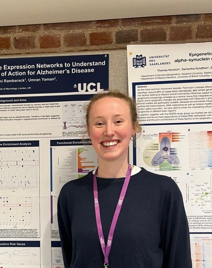 Smiling presenter in a dark top and purple lanyard stands before research posters at a conference.