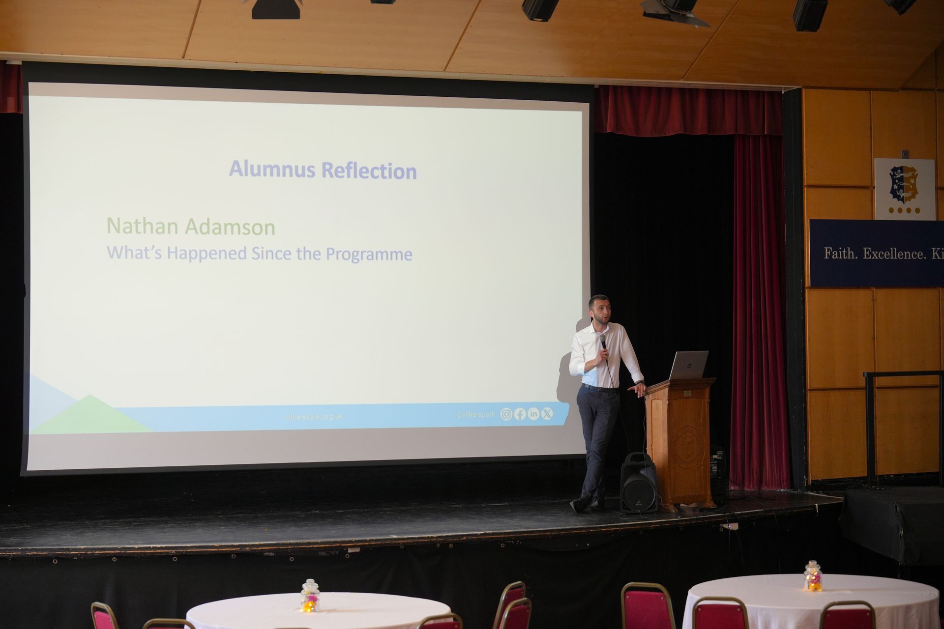Presenter beside projection screen in a theater, speaking on stage during a lecture on 