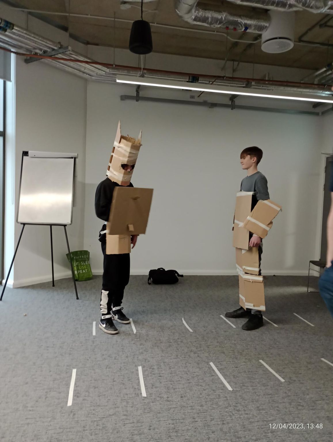 Two people in cardboard costumes standing in a studio with a flip chart and taped floor lines
