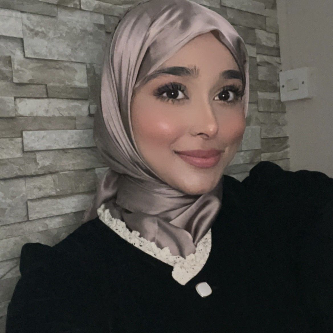 Smiling person in a silver hijab and black top against a gray stone wall.