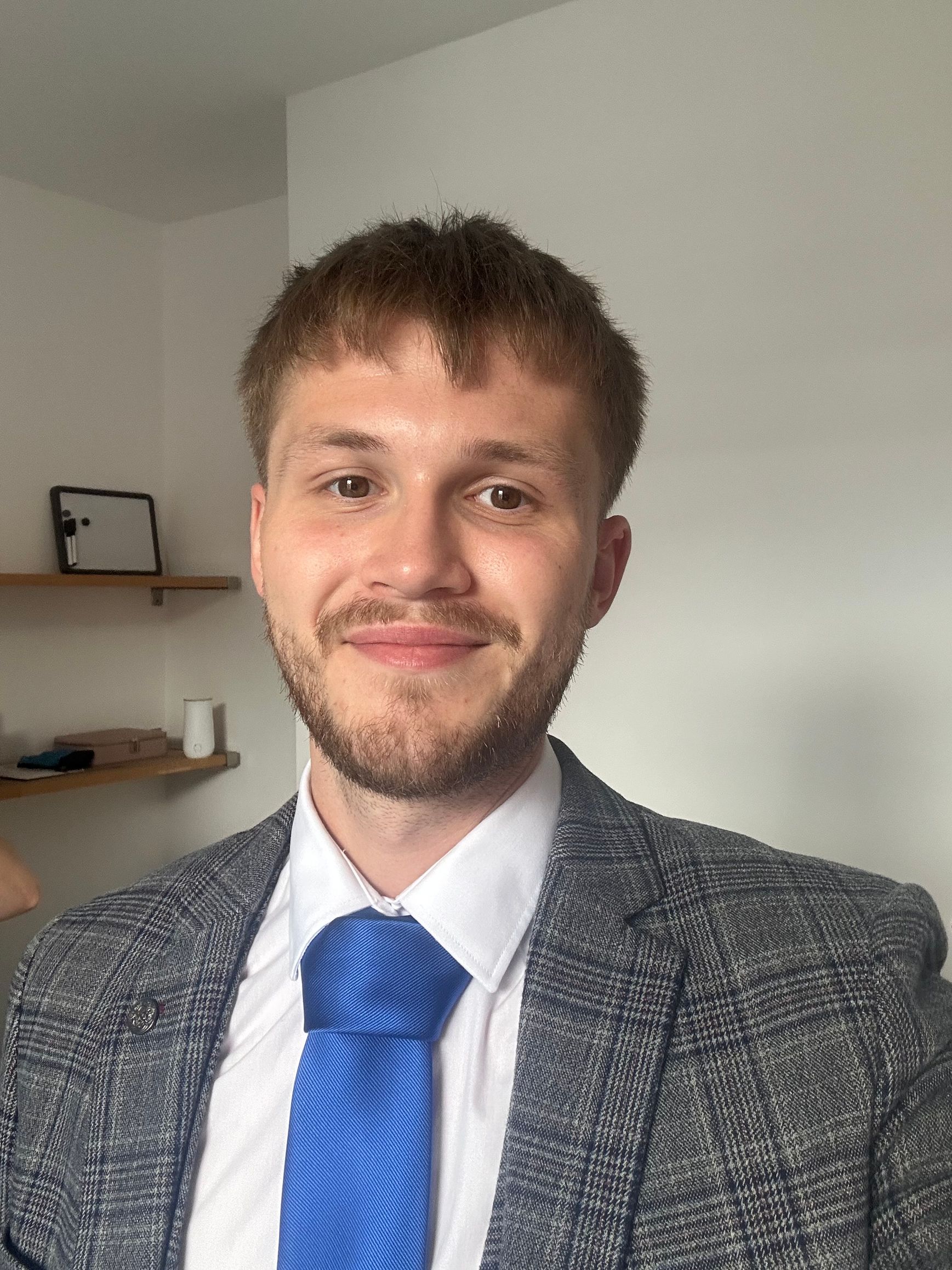 Smiling person in a gray blazer and blue tie standing indoors against a white wall.