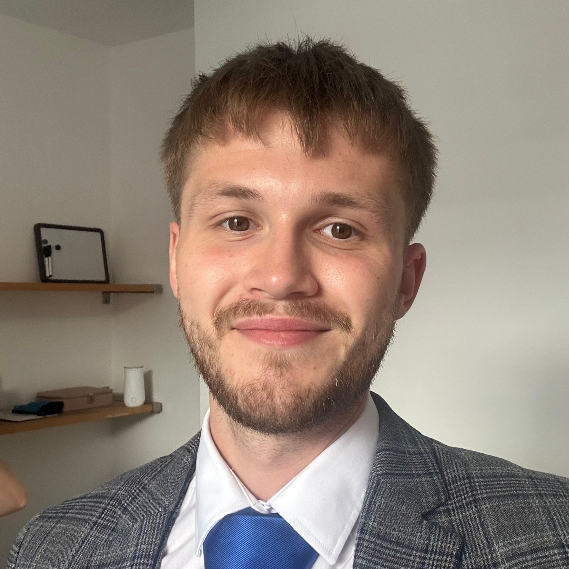 Man in a blue tie and gray plaid jacket smiling indoors beside wall shelves