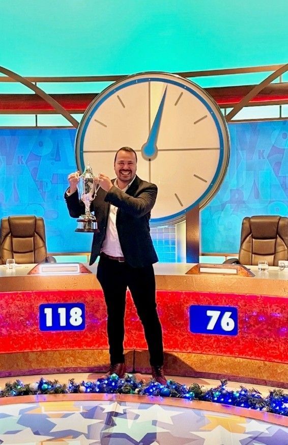 Man holding a trophy on a game show stage with a large clock and scoreboards reading 118 and 76