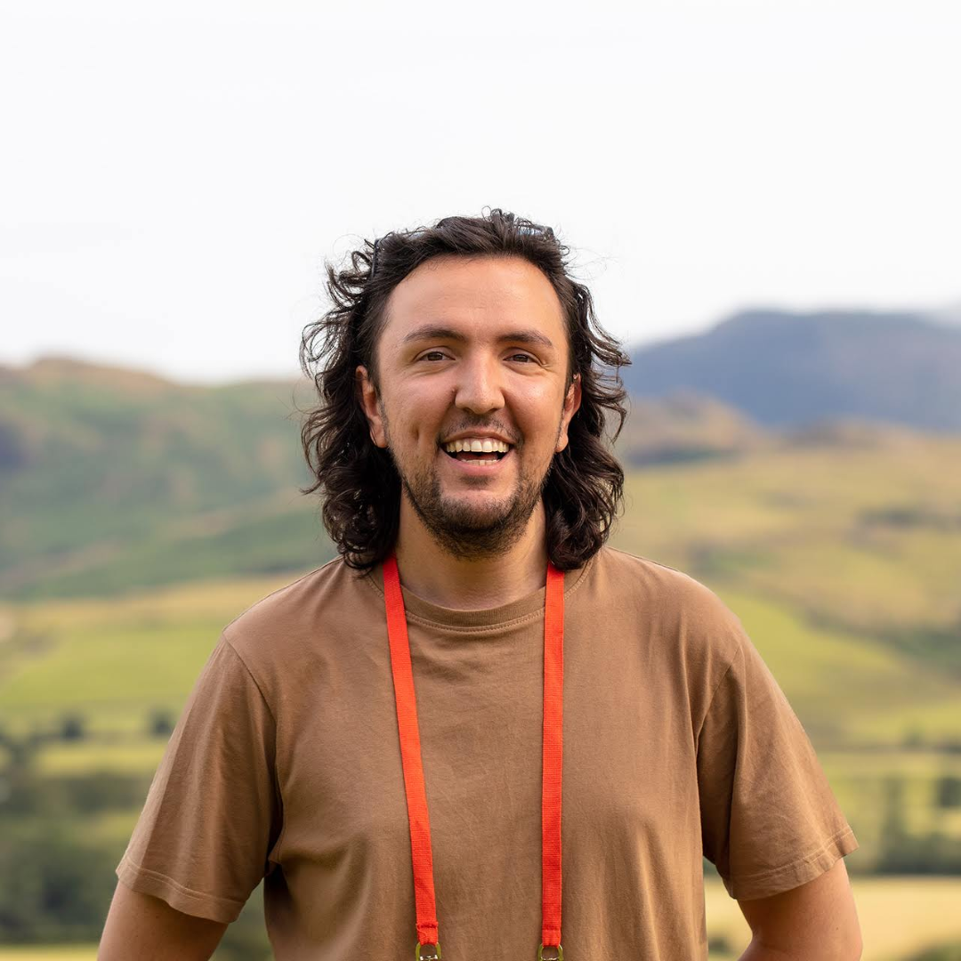 Smiling person with red lanyard in a brown shirt standing outdoors with hills in the background