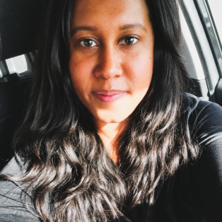 Close-up selfie of a person with long wavy hair in a car, wearing a dark top and looking at the camera